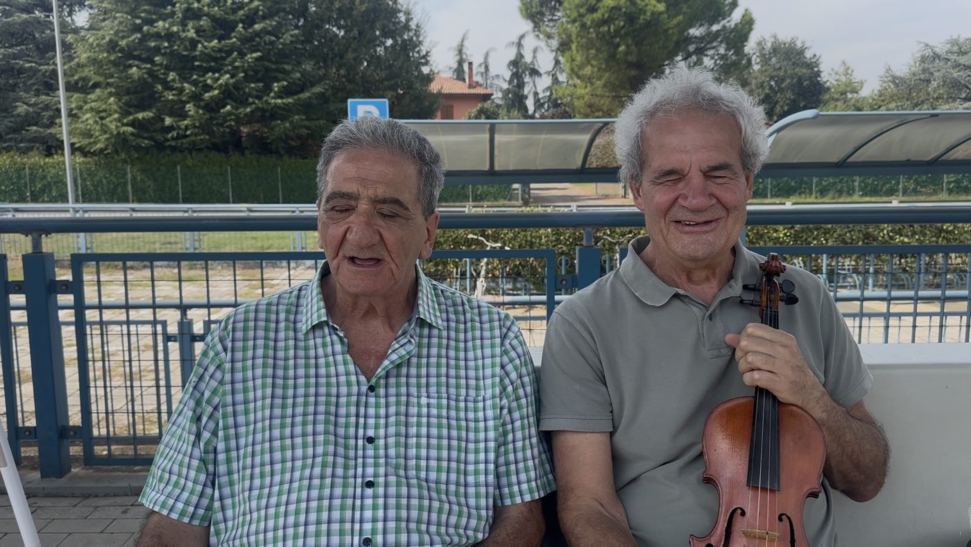 Maurizio e Paolo seduti sulla panchina della Stazione di Casteldebole, Paolo tiene il violino
